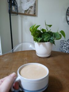 At a dining table with a plant on it; a cup of coffee in hand.